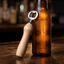 A wooden-handled bottle opener with a silver metal opener, resting against an amber glass beer bottle on a dark wooden table.