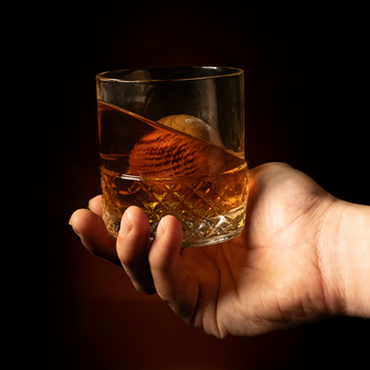 Hand holding a glass of whiskey with an oak ice sphere resting in it.